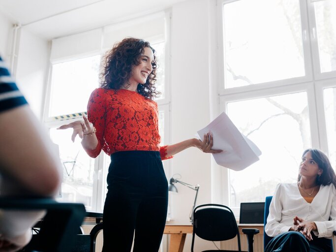 Eine Frau steht im Büro und hält eine Präsentation vor ihren Kolleg*innen  | © Getty Images / Hinterhaus Productions