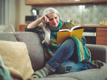 Frau in mittlerem Alter sitzt auf der Couch und liest ein Buch | © Getty Images/Eva-Katalin