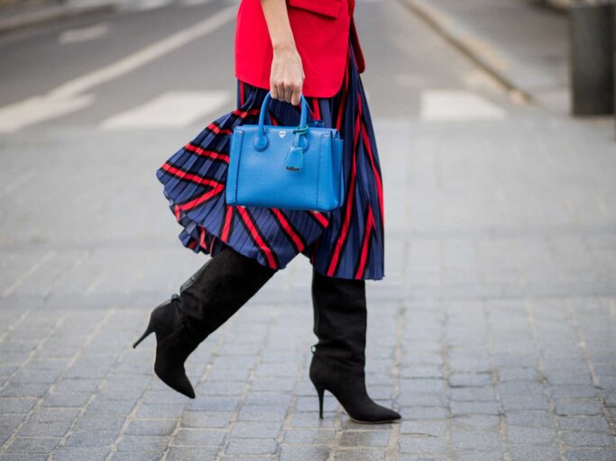 Nahaufnahme einer Frau, die einen rot-blauen Faltenrock trägt, schwarze Stiefel und eine blaue Tasche | © Getty Images/Christian Vierig