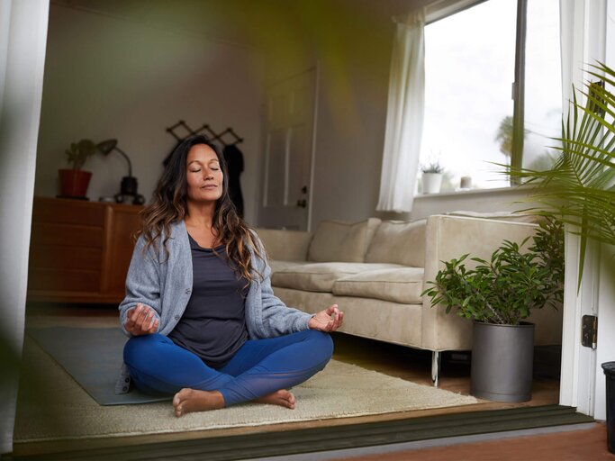 Reife Frau meditiert beim Yoga-Üben in ihrem Wohnzimmer. | © iStock.com / Goodboy Picture Company