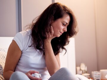 Frau sitzt auf dem Bett und hält sich den Hals vor Schmerzen. | © iStock.com / dusanpetkovic